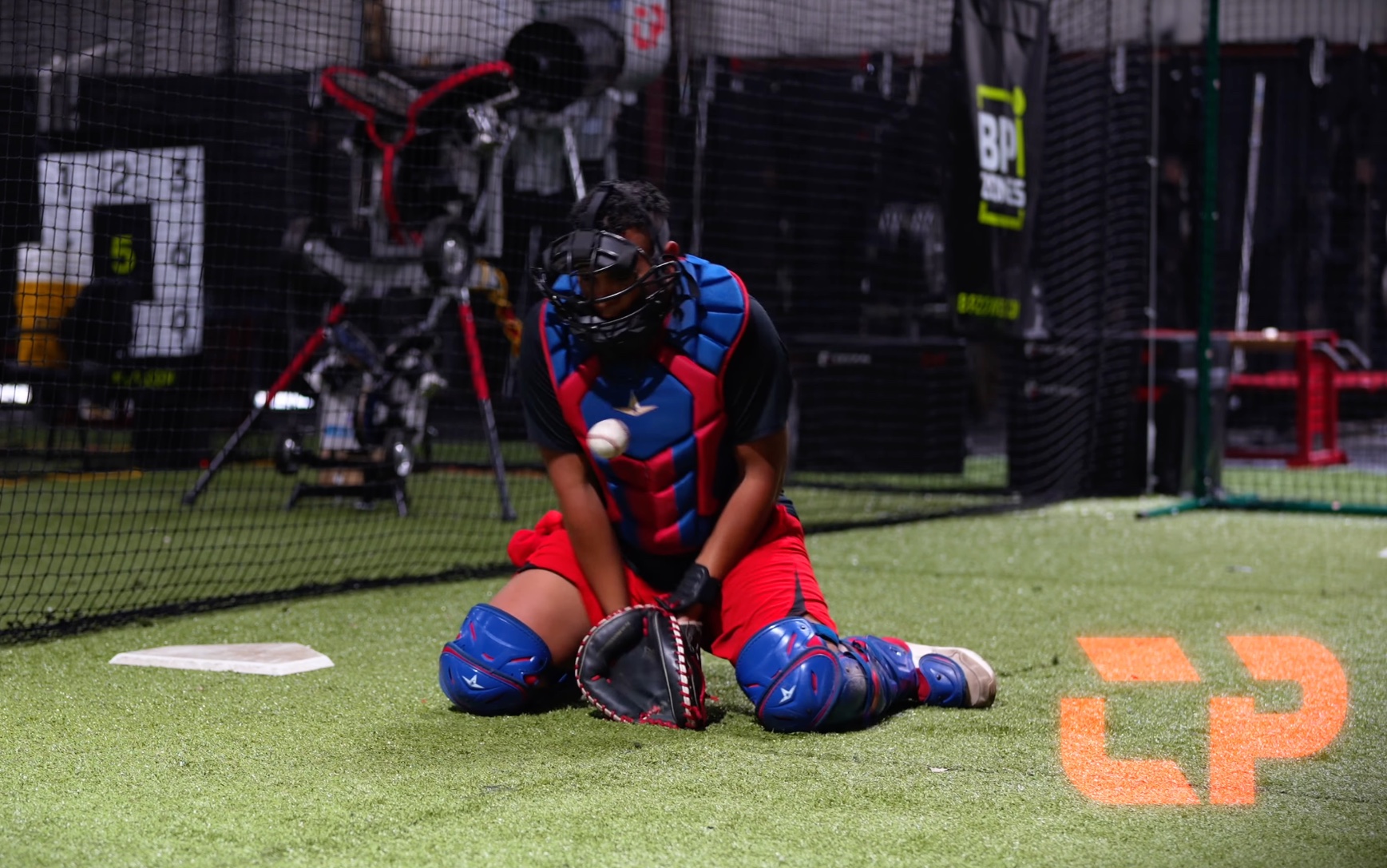 Catcher in blue and red gear keeping a blocked baseball in front on indoor turf